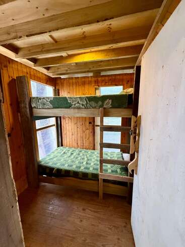 Esse quarto aconchegante de madeira tem beliches com cobertores verdes xadrez e florais, paredes de madeira, janelas grandes, vigas expostas no teto e uma escada simples de madeira.