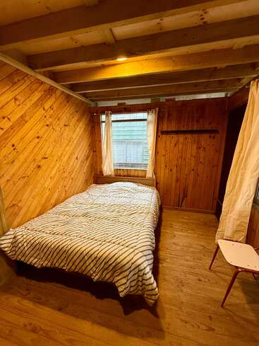 Esse quarto aconchegante de madeira apresenta uma cama simples com um edredom listrado, duas janelas com cortinas, paredes com painéis de madeira, vigas expostas e uma pequena mesa lateral branca.