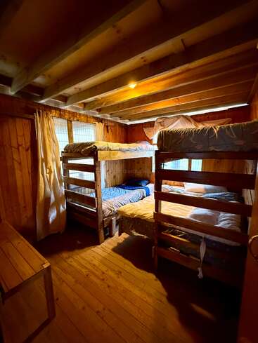 Esse quarto aconchegante de madeira tem dois conjuntos de beliches, iluminação suave, vigas expostas, paredes e piso de madeira, janelas com cortinas, criando uma atmosfera de cabana rústica e aconchegante.