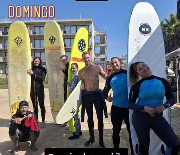 Un groupe de six personnes pose avec des planches de surf sur une plage ensoleillée, portant des combinaisons de plongée et souriant. Le mot "DOMINGO" apparaît en haut de l'image.