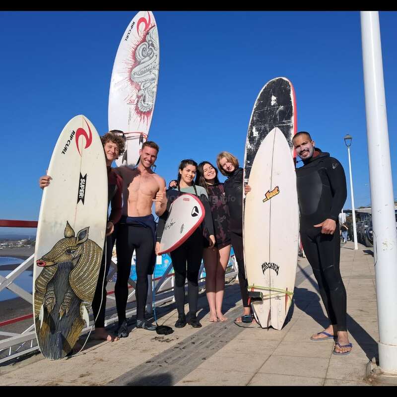 Six amis tenant des planches de surf sourient sur une promenade ensoleillée au bord de l'océan. Ils sont vêtus de combinaisons et de maillots de bain, prêts à s'amuser à surfer ensemble.