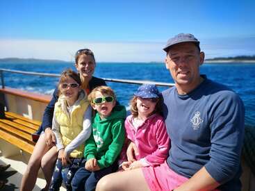 Das Bild zeigt eine fünfköpfige Familie, die auf einem Boot sitzt, mit einem Gewässer und einem blauen Himmel im Hintergrund, und gemeinsam einen sonnigen Tag genießt.