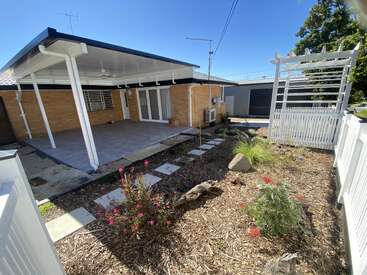 A neat backyard features a covered patio, stepping stones, mulched garden beds with flowering plants, a white trellis, brick house, and a grey shed under blue sky.