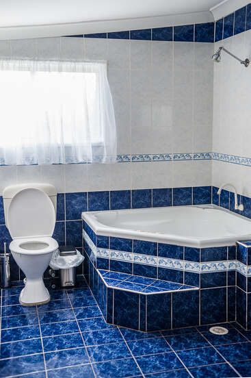 Dieses Badezimmer hat blaue und weiße Fliesen, eine Badewanne mit Stufe, eine Toilette, ein kleines Fenster mit einem durchsichtigen Vorhang und einen Mülleimer neben der Toilette.