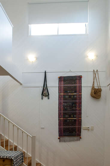 Ein helles, weißes Treppenhaus hat ein Fenster, zwei Wandleuchten, einen gemusterten Wandteppich, zwei gewebte Taschen und eine Treppe mit einem karierten Tuch drauf.