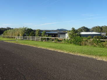 The image depicts a serene rural scene with a road, fence, and buildings, set against a backdrop of trees and a clear blue sky on a sunny day.