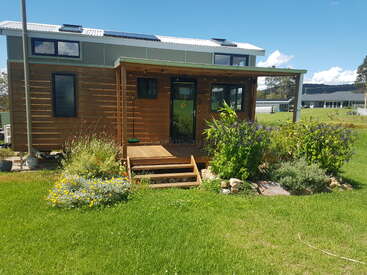 The image depicts a small, wooden house with a porch and stairs, surrounded by lush green grass and vibrant flowers, set against a serene blue sky.