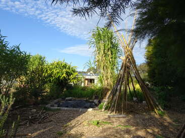 The image depicts a serene garden scene with a teepee structure, surrounded by lush greenery and a small pond, set against a bright blue sky with scattered clouds.