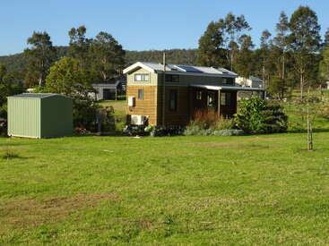 The image depicts a serene rural scene featuring a small, two-story house with a shed and lush greenery, set against a backdrop of trees and a clear blue sky.
