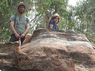 A man wearing a hat and a child sit on top of a large rock formation, surrounded by green trees, enjoying an outdoor adventure together.