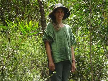 A woman stands in lush green foliage, wearing a wide-brimmed hat and loose green clothing, smiling and enjoying the natural outdoor, sunlit environment around her.