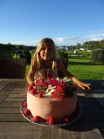 A smiling young girl sits outdoors at a wooden table with a large pink birthday cake decorated with flowers and raspberries, surrounded by green grass and houses.
