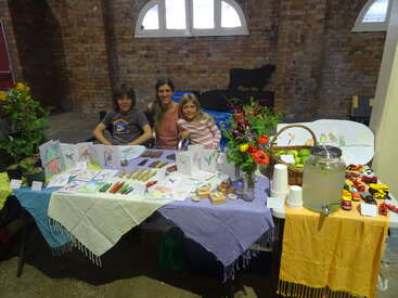 Three smiling people sit behind a colorful market stall displaying handmade crafts, cards, flowers, lemonade, and toy cars, all set in a rustic brick building.