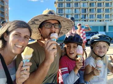Das Bild zeigt eine vierköpfige Familie, die an einem sonnigen Tag Eiswaffeln genießt, mit einem großen Gebäude und Autos im Hintergrund.