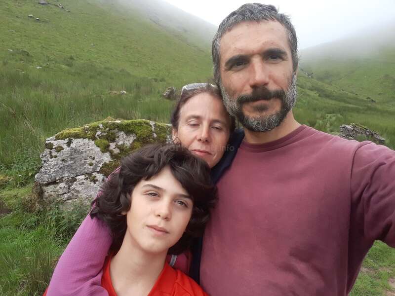 Eine dreiköpfige Familie posiert eng zusammen im Freien. Grüne Hügel und nebliges Wetter schaffen eine ruhige, friedliche Kulisse. Sie wirken entspannt und genießen die Zeit in der Natur.
