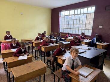 Un aula alegre con alumnos jóvenes sentados en pupitres de madera, vestidos con uniforme. La luz del sol entra a raudales por los grandes ventanales. Todos parecen concentrados, con cuadernos y bolígrafos sobre los pupitres.