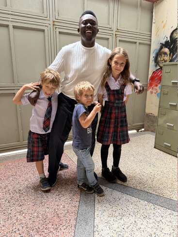 Un hombre sonriente, vestido con un jersey blanco, posa junto a tres niños —dos de ellos con uniforme escolar— en el interior de una habitación, con una pared pintada y unas taquillas como fondo.
