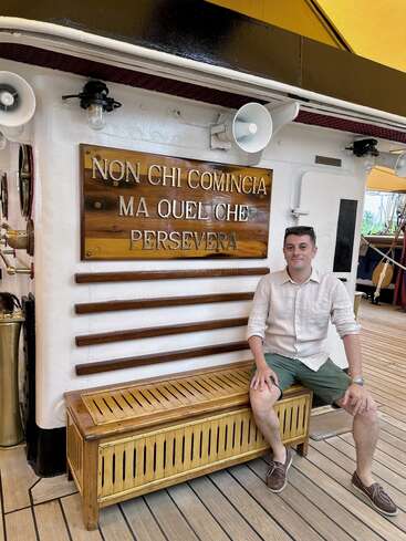 A man sits on a wooden bench aboard a ship, smiling. Behind him is an Italian sign reading, “Non chi comincia ma quel che persevera.”