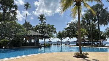A tropical resort pool surrounded by palm trees, sun loungers, and shaded cabanas. The blue sky and distant mountains create a peaceful, vacation-like atmosphere.
