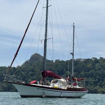 Ein weißes Segelboot mit kastanienbraunen Akzenten schwimmt friedlich auf ruhigem Wasser, umgeben von üppig grünen Hügeln und unter einem leicht bewölkten Himmel. Zwei Menschen sind an Bord.
