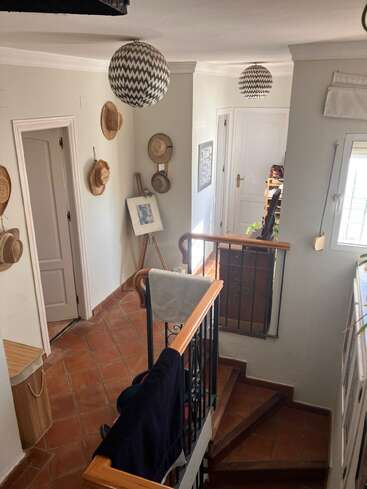 Das ist ein gemütliches Treppenhaus mit Terrakottafliesen, Lampen mit Zickzackmuster, dekorativen Hüten an der Wand, einem gerahmten Bild und natürlichem Sonnenlicht, das reinkommt.
