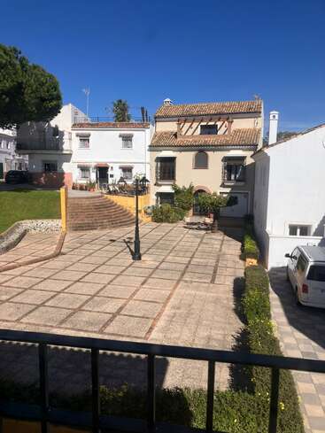 Das Bild zeigt einen sonnigen Innenhof mit gefliesten Böden, der von Büschen umrandet ist und von charmanten Häusern im mediterranen Stil mit Ziegeldächern und einem strahlend blauen Himmel umgeben ist.