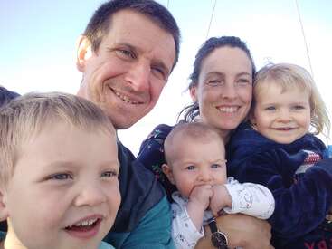 Eine fröhliche fünfköpfige Familie posiert eng beieinander und lächelt in die Kamera. Die Eltern halten ihre drei kleinen Kinder im Arm und das helle Tageslicht beleuchtet ihre glücklichen Gesichter.