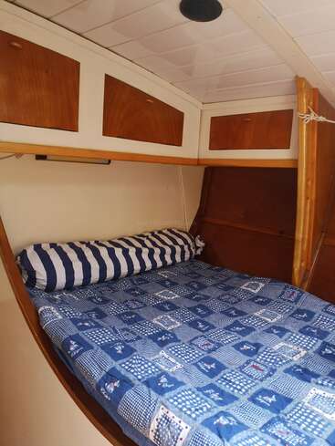 Dieses Bild zeigt ein gemütliches Kajütbett mit einer blauen Patchworkdecke, gestreiften zylindrischen Kissen, Holzschränken darüber, weißen Deckenpaneelen und hellen Holzakzenten.
