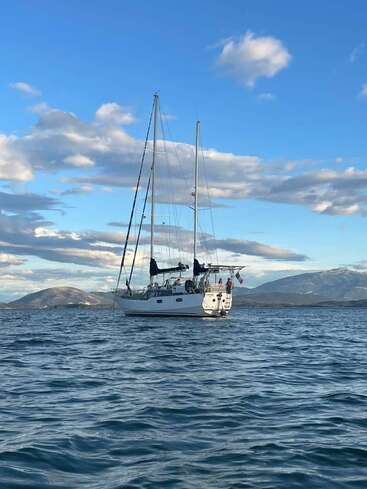 A white sailboat floats calmly on blue water under a bright sky with fluffy clouds. Mountains rise in the background, creating a serene, picturesque scene.