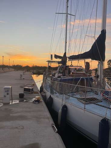 A sailboat is docked at a quiet marina during sunset. The sky is colorful, and marina equipment is scattered along the concrete pier, evoking tranquility.