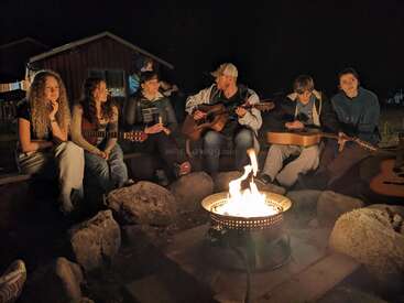 Un groupe de personnes est assis autour d'un feu de camp, la nuit, jouant de la guitare, discutant et profitant ensemble de la musique. L'ambiance est chaleureuse, rustique et détendue.