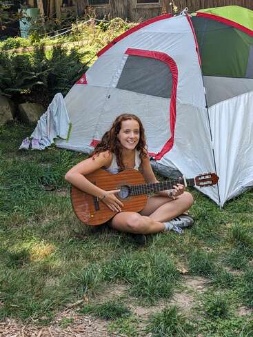 Une jeune fille aux cheveux bouclés est assise en tailleur sur l'herbe ; elle sourit et joue de la guitare devant une tente de camping, profitant d'une journée ensoleillée en plein air.