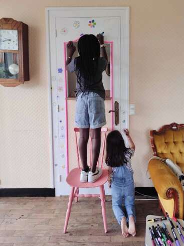 Deux enfants dessinent avec enthousiasme des fleurs et des formes colorées sur une porte blanche. L'un est debout sur une chaise rose, tandis que l'autre est à genoux, tous deux complètement absorbés par leur activité.