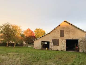 Une grange rustique aux portes ouvertes se dresse dans une cour herbeuse, entourée d'arbres aux couleurs automnales, sous un ciel pastel en début de soirée.