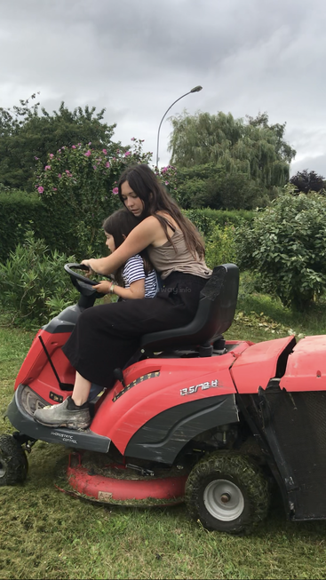 Une femme et un enfant conduisent ensemble une tondeuse à gazon rouge dans un jardin, entourés d'une végétation luxuriante et de fleurs, sous un ciel nuageux et couvert.