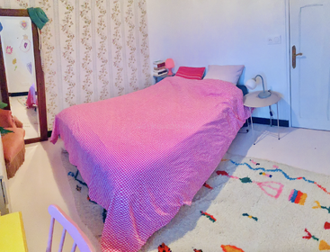 Cette chambre confortable est équipée d'un couvre-lit à carreaux roses, d'un tapis coloré, de tables de chevet avec lampes, d'une pile de livres, d'un papier peint à fleurs et d'un miroir en pied.