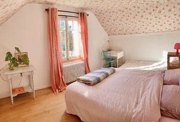 Cette chambre confortable est dotée d'un couvre-lit rose, d'un plafond fleuri, d'un parquet, d'une fenêtre avec des rideaux roses, d'un petit bureau, de plantes et d'une lumière naturelle chaleureuse.