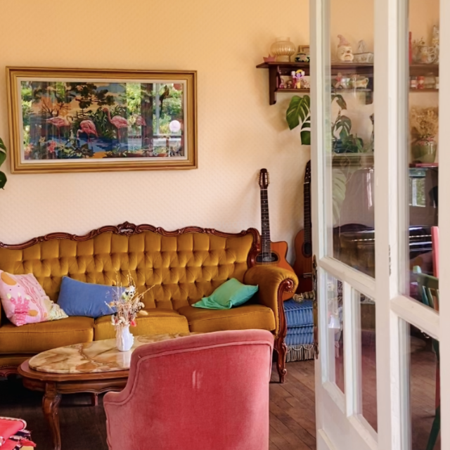 Un salon vintage confortable comprend un canapé capitonné jaune moutarde, des coussins colorés, une table basse en marbre, une guitare, des étagères, des plantes, des œuvres d'art et un éclairage chaleureux.