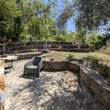 Un jardin paisible avec des terrasses en pierre, une chaise en plastique, une table et diverses plantes en pot. La lumière du soleil filtre à travers les arbres à la verdure luxuriante, projetant des ombres tachetées sur le sol.