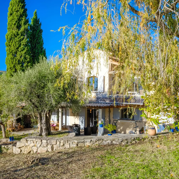Das Bild zeigt eine ruhige Szene eines Hauses mit einem großen Garten, in dem ein Baum und ein Zaun stehen, vor dem Hintergrund eines klaren Himmels und eines Gewässers.