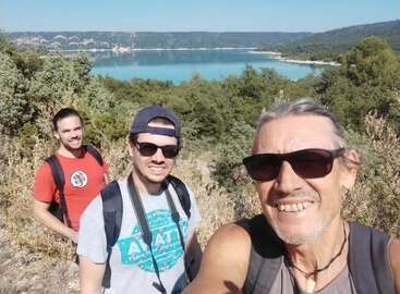 La imagen muestra a tres hombres tomándose un selfie frente a un lago sereno, rodeado de árboles y colinas, en un día soleado.