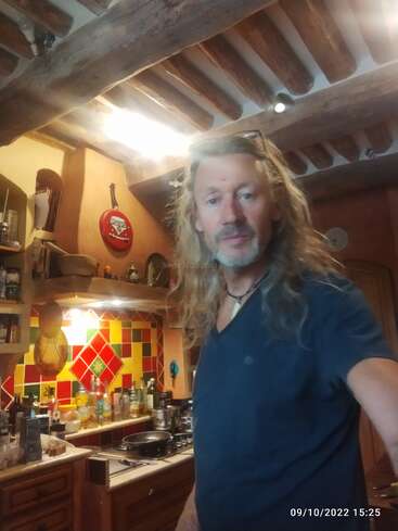 La imagen muestra a un hombre de pelo largo de pie en una cocina, rodeado de diversos utensilios e ingredientes para cocinar, con un colorido salpicadero y techo de madera.
