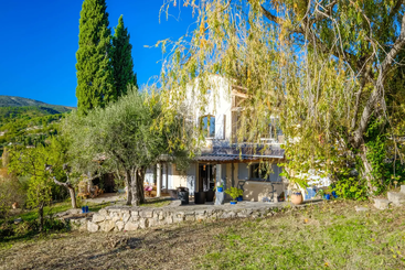 La imagen representa una serena escena de una casa con un gran patio, un árbol y una valla, con un cielo despejado y una masa de agua como telón de fondo.