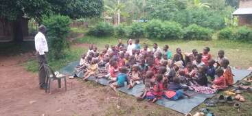 Eine Gruppe von Kindern sitzt im Freien auf Matten und hört aufmerksam einem Mann zu, der vor ihnen steht. Die Schuhe liegen neben den Matten auf dem Boden.
