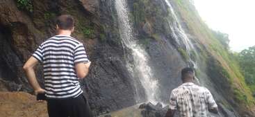 Zwei Männer stehen am Fuße eines Wasserfalls an einem felsigen, bewaldeten Berghang. Beide scheinen die Natur zu beobachten, umgeben von üppigem Grün und Nebel.