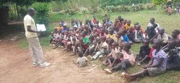 Ein Mann steht mit einem Buch in der Hand da und unterrichtet eine große Gruppe von Kindern, die aufmerksam auf dem Boden im Freien sitzen, umgeben von Grün und einer natürlichen Umgebung.