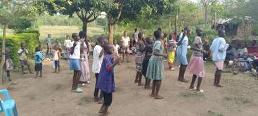 A group of children are dancing and playing outside under trees. Other children and adults watch, sitting nearby. The atmosphere is lively, joyful, and communal.