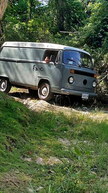 Ein klassischer grauer Van fährt durch einen unwegsamen, grasbewachsenen Offroad-Weg, der von dichtem grünen Wald umgeben ist. Man sieht eine Person, die ihren Arm nach draußen streckt.
