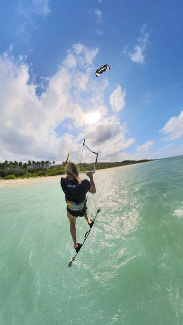 Eine Person fährt mit dem Kiteboard über kristallklares türkisfarbenes Wasser in der Nähe eines Sandstrandes, unter einem hellen, sonnigen Himmel mit vereinzelten Wolken und üppigem Grün in der Nähe.