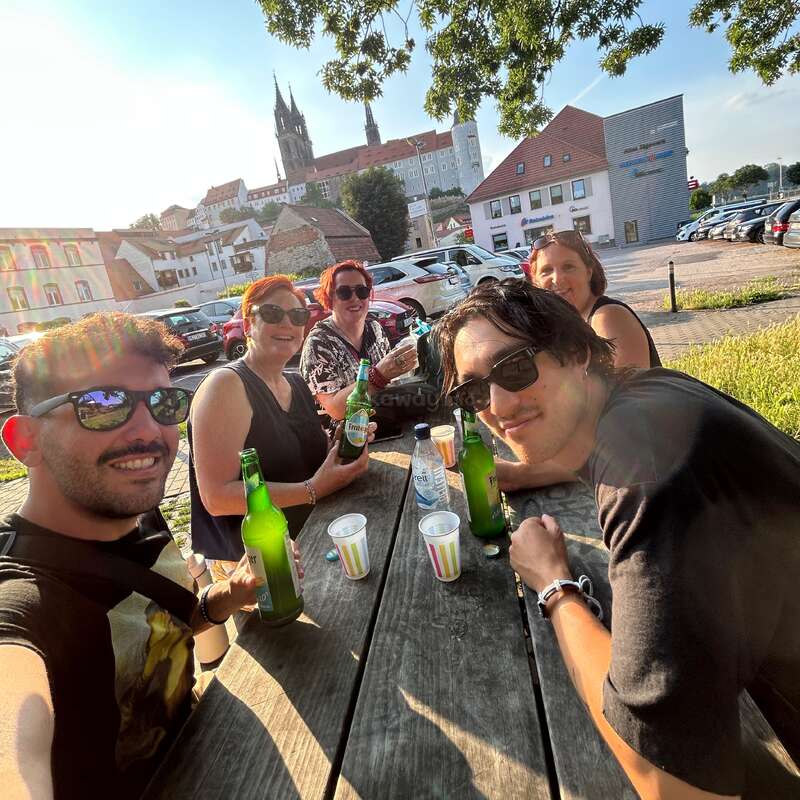 Cinco amigos sentados em uma mesa de piquenique de madeira ao ar livre, desfrutando de bebidas e sorrisos sob um céu ensolarado. Edifícios históricos e uma torre de igreja estão ao fundo.